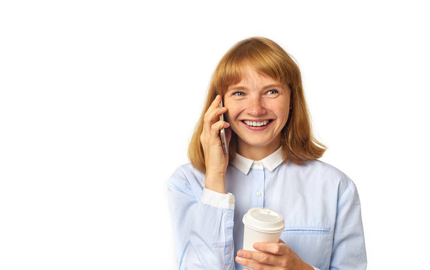 happy female model with ginger hair and freckles dressed in blue shirt talking on smartphone and holding coffee cup  while posing isolated on white background - Photo, image