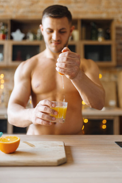 Man with naked body making orange juice on the kitchen. Nude male person preparing breakfast at home, food preparation without clothes - Photo, image