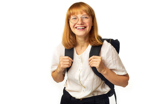 happy redheaded woman with freckles wearing glasses and white t-shirt with big black backpack laughing while looking at camera isolated on white background - Foto, Imagen