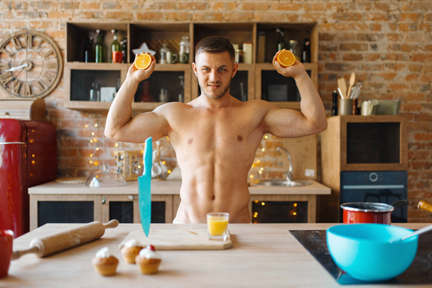 Facet z nagim ciałem robi sok pomarańczowy w kuchni. Nagi mężczyzna przygotowuje śniadanie w domu, przygotowuje jedzenie bez ubrania - Zdjęcie, obraz