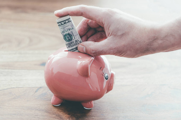 zbliżenie męski ręka wprowadzenie składany dolar banknot w różowy świnka bank na drewniany stół - Zdjęcie, obraz