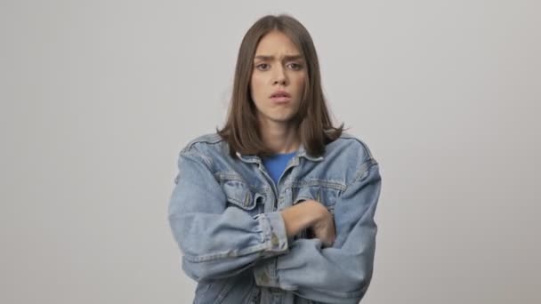Upset young brunette woman in denim jacket crossing her arms and becoming offended while looking at the camera over gray background isolated - Imágenes, Vídeo
