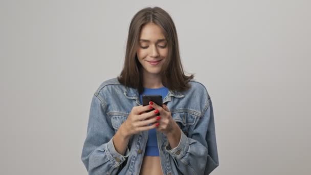 Happy young brunette woman in denim jacket smiling and laughing while using smartphone over gray background isolated - Кадри, відео