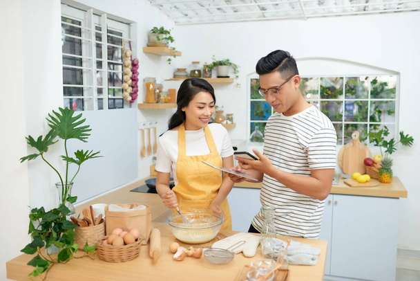 Молодий азіатський чоловік і жінка разом готують торт і хліб з яйцем, дивлячись меню з таблетки в борошні щасливий відпочинок вдома
 - Фото, зображення