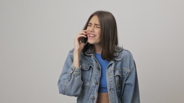 Displeased young brunette woman in denim jacket arguing and becoming angry while talking on smartphone over gray background isolated - Video, Çekim
