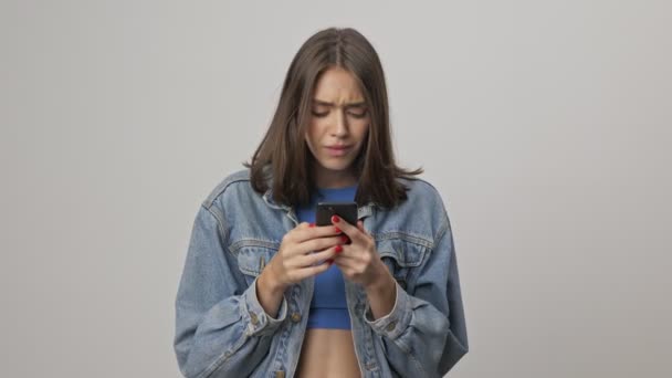Unhappy young brunette woman in denim jacket becoming displeased and feeling disgusted while using smartphone over gray background isolated - Video, Çekim