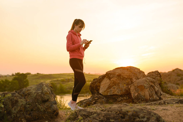 Woman Runner Resting after Workout, Using Smartphone and Listening to Music at Sunset on the Rock. Sports Concept. - Φωτογραφία, εικόνα