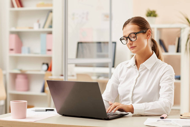 Joven secretaria en anteojos y camisa blanca sentada a la mesa y trabajando online en laptop en oficina - Foto, imagen