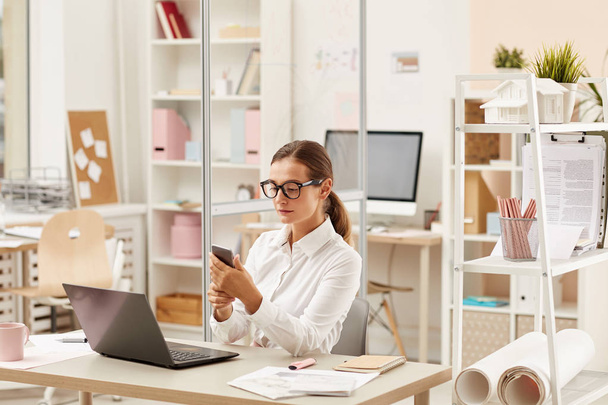 Jonge zakenvrouw in bril zit aan tafel voor laptop en na te denken over nieuwe ideeën tijdens het werken op kantoor  - Foto, afbeelding