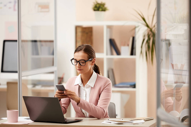 Vážná podnikatelka v brýlích pomocí mobilního telefonu a komunikace on-line při práci u stolu s notebookem v kanceláři - Fotografie, Obrázek