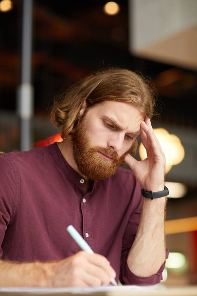 Serious red haired businessman sitting at the table and writing in document preparing for the meeting - 写真・画像