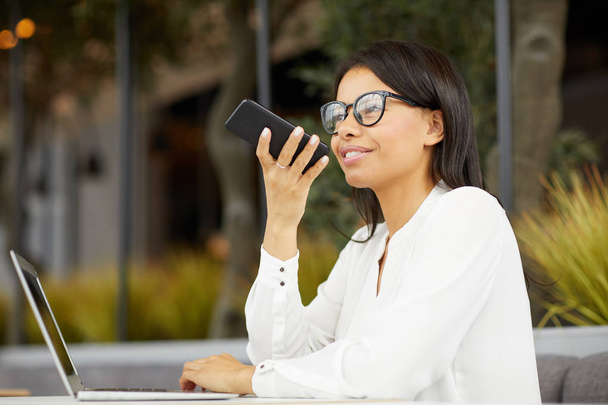 Young beautiful businesswoman in eyeglasses smiling and recording audio message on mobile phone while sitting at the table with laptop outdoors - Photo, image