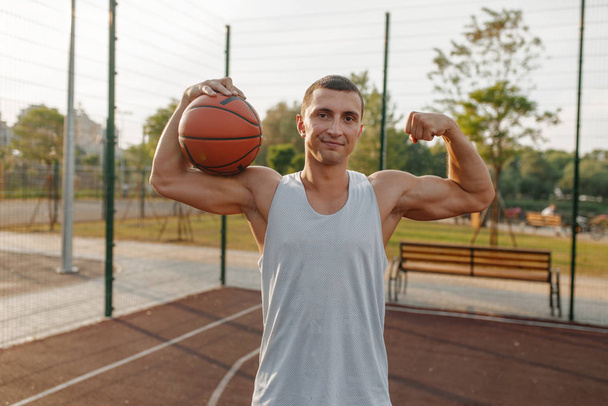 Άρρεν μπασκετμπολίστας με μπάλα δείχνει τους μυς του σε υπαίθριο γήπεδο, μπροστινή άποψη. Άνδρας αθλητής σε αθλητικά είδη στην προπόνηση streetball, θερινό στάδιο - Φωτογραφία, εικόνα