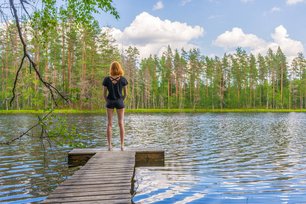 Cute ginger girl standing on wooden planked footway in summer day against beautiful landscape of northern lake and forest. Travelling and discovering distant places of Earth. Karelia, Russia - Фото, зображення