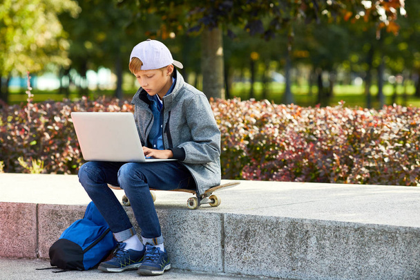 Αγόρι σε casual ρούχα κάθεται σε skateboard και πληκτρολογώντας σε φορητό υπολογιστή σε εξωτερικούς χώρους προετοιμασία για την εργασία - Φωτογραφία, εικόνα