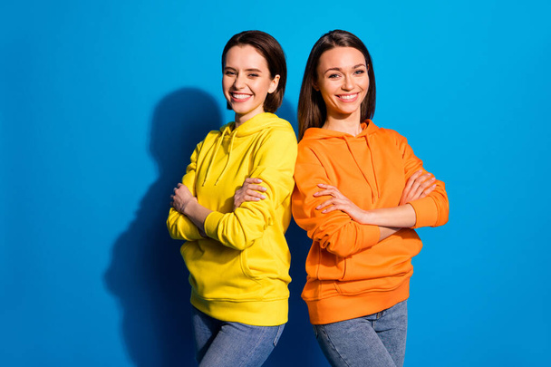 Photo of pretty two girlfriends lady standing back-to-back in good mood crossing arms wear casual bright hoodies and jeans isolated blue color background - Photo, image