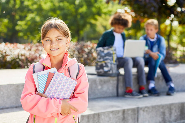Portrait d'écolière mignonne tenant des livres et souriant à la caméra tout en se tenant dans le parc avec des camarades de classe en arrière-plan - Photo, image