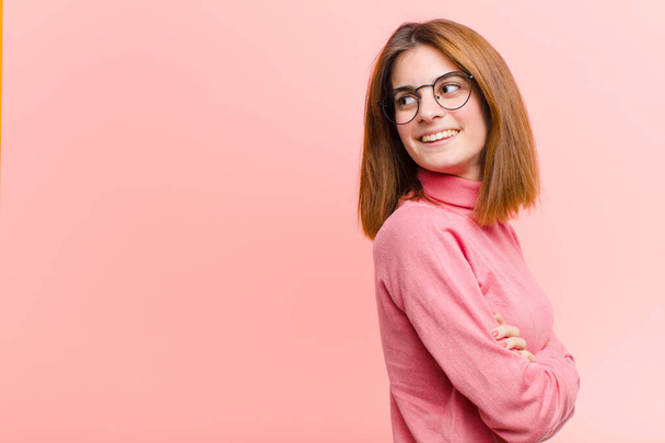 young pretty woman smiling gleefully, feeling happy, satisfied and relaxed, with crossed arms and looking to the side against pink background - Photo, image