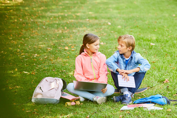 Школьница сидит на зеленой траве с ноутбуком и разговаривает с другом, готовясь к урокам вместе перед школой - Фото, изображение