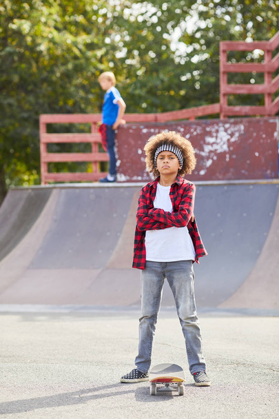 Portrait of African boy with curly hair standing with arms crossed and looking at camera while riding on skateboard outdoors - Foto, Bild