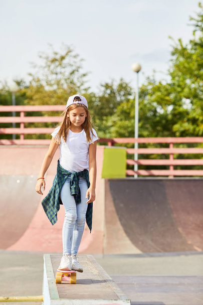 Girl in casual clothing standing on skateboard and learning to ride in the skateboard park outdoors - 写真・画像