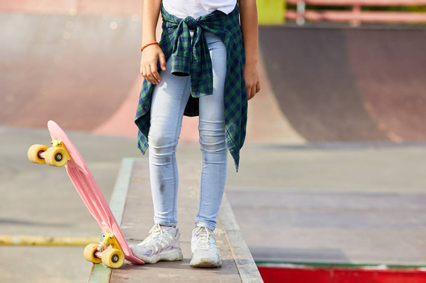 Close-up de menina em jeans de pé e segurando com pé skate enquanto cavalga no parque de skate ao ar livre - Foto, Imagem