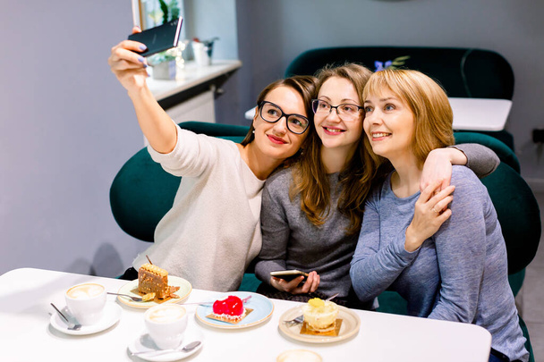 three young beautiful women sitting at cafe indoors, happy, having fun, smiling, looking at smartphone, taking selfie photo, flirty - Foto, Imagen