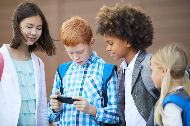 Gruppe von Schülern steht und schaut Klassenkameraden beim Spielen auf dem Handy zu  - Foto, Bild