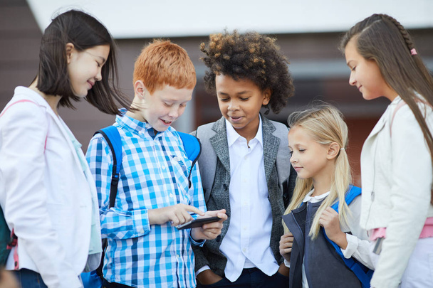 Grupo de compañeros de clase con mochilas de pie juntos y mirando al niño con el teléfono móvil al aire libre
 - Foto, Imagen