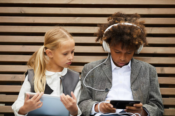 African schoolboy wearing headphones and listening to audio lesson on mobile phone together with schoolgirl sitting on the bench outdoors - Фото, изображение