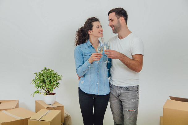 Shot of charming copule standing with glasses of champange, celebrating their move into new apartment, smiling, looking at one another with love in eyes. - 写真・画像