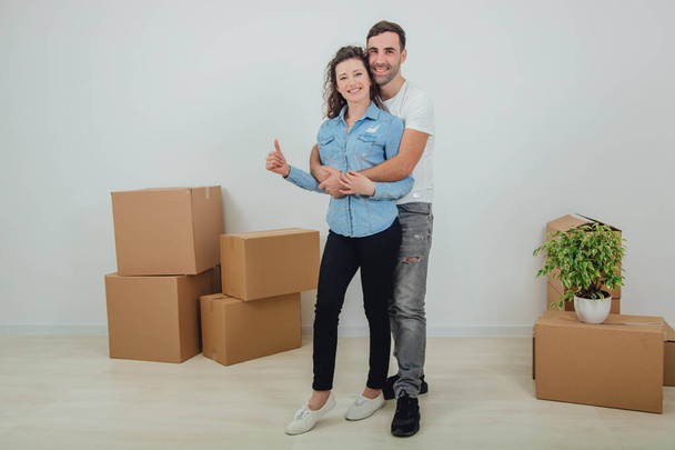 Couple moving into their new house, husband is hugging his wife, looking at the camera, smiling. Wife is giving thumb up. Room full of carton boxes. - Fotografie, Obrázek