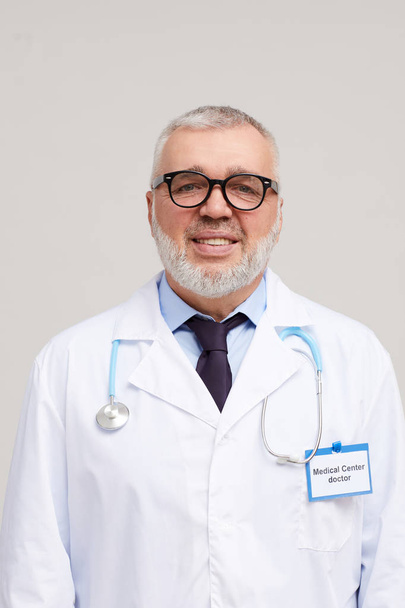 Retrato de médico maduro en anteojos y en bata blanca sonriendo a la cámara sobre fondo blanco
  - Foto, imagen