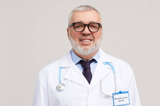 Portret dojrzałego lekarza w okularach i w białym płaszczu uśmiechniętym do kamery na białym tle  - Zdjęcie, obraz