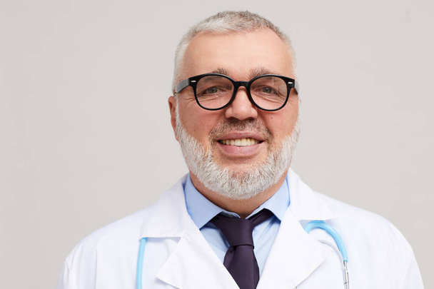 Porträt eines reifen Arztes mit Brille und weißem Mantel, der auf weißem Hintergrund in die Kamera lächelt  - Foto, Bild