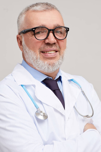 Retrato de médico maduro en anteojos y en bata blanca sonriendo a la cámara sobre fondo blanco
  - Foto, Imagen