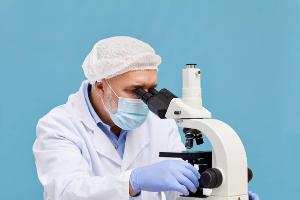 Médico maduro en máscara protectora y en sombrero examinando la muestra en el microscopio sobre fondo azul
  - Foto, Imagen