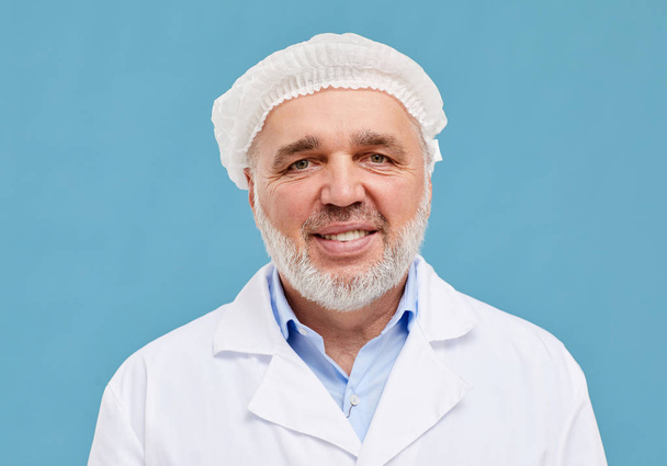 Portrait of mature successful surgeon in hat and white coat smiling at camera on blue background - Fotó, kép