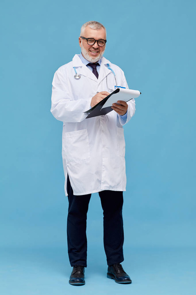 Portrait of mature doctor in white coat in eyeglasses holding folder and smiling at camera over blue background  - Foto, afbeelding
