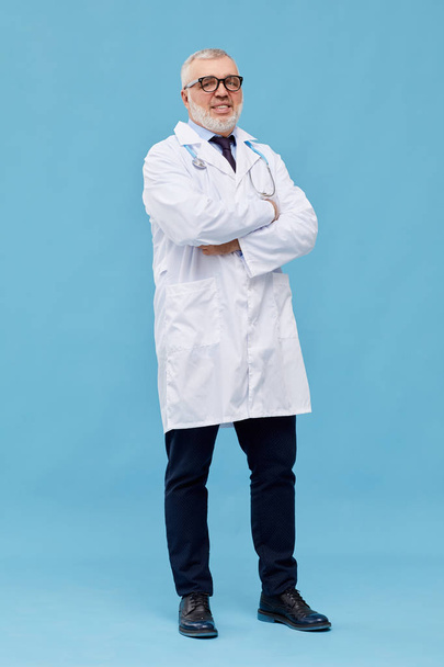 Férfi érett orvos portréja szemüvegben és fehér kabátban, keresztbe tett karokkal és a kamerát kék alapon nézve - Fotó, kép