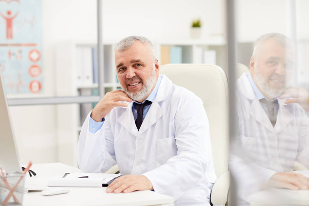 Портрет успішного старшого лікаря в білому пальто, посміхаючись на камеру, сидячи за столом в офісі
  - Фото, зображення