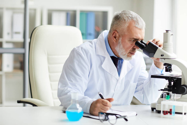 Σοβαρή ώριμη επιστήμονας επικεντρώνεται στην εργασία κάθεται στο τραπέζι και κοιτάζοντας μέσα από το μικροσκόπιο στο γραφείο  - Φωτογραφία, εικόνα