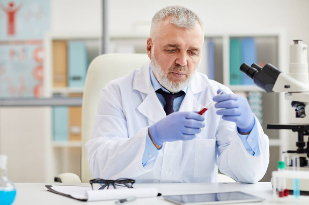 Ανώτερος γιατρός με προστατευτικά γάντια κάθεται στο τραπέζι εξετάζοντας σωλήνες κατά τη διάρκεια της ιατρικής εξέτασης  - Φωτογραφία, εικόνα