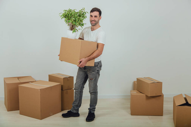 Handsome young man moving into new house, standing, holding carton box and flowerpot, smiling, surrounded by boxes. - 写真・画像