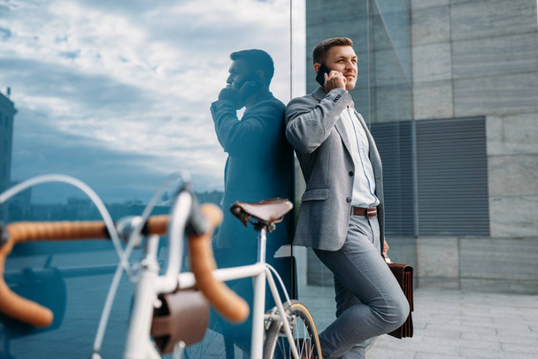 Businessman in suit talking by mobile phone at the bicycle in downtown. Business person riding on eco transport on the street - Fotoğraf, Görsel