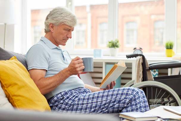 Uomo anziano in abiti domestici appoggiato sul divano con tazza di caffè e libro di lettura in soggiorno a casa
 - Foto, immagini