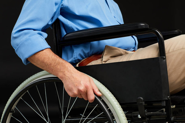Primer plano del paciente mayor sosteniendo la rueda de la silla de ruedas mientras está sentado sobre un fondo negro
 - Foto, imagen