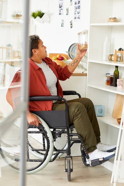 Αφρικανική ανάπηρος γυναίκα κάθεται σε αναπηρική καρέκλα κοντά στα ράφια και κοιτάζοντας το προϊόν στο χέρι στην κουζίνα - Φωτογραφία, εικόνα