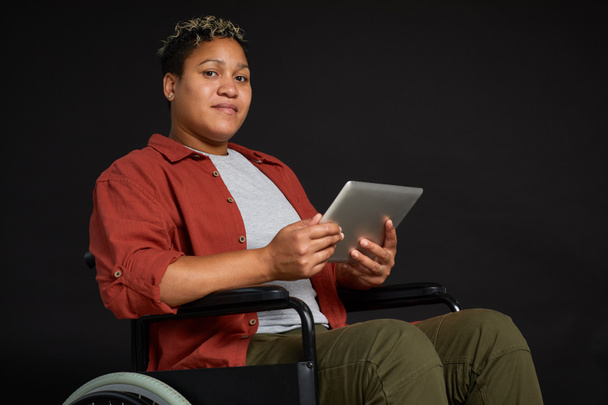 Retrato de una joven africana discapacitada en silla de ruedas mirando la cámara mientras usa la tableta PC sobre fondo negro
 - Foto, Imagen