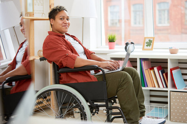 Porträt einer jungen Frau mit Behinderung, die im Rollstuhl sitzt und in die Kamera schaut, während sie zu Hause ihren Laptop benutzt - Foto, Bild